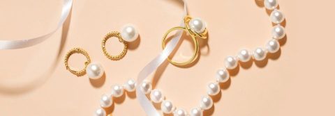 Flat lay of gold pearl jewelry — a bracelet, ring, drop earrings and pendant charm — styled with a white ribbon on a soft peach background.