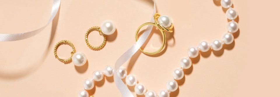 Flat lay of gold pearl jewelry — a bracelet, ring, drop earrings and pendant charm — styled with a white ribbon on a soft peach background.