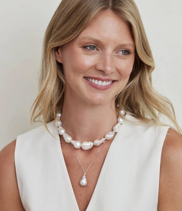 Woman smiling in a cream top, wearing a layered look of a large baroque pearl strand necklace and a delicate chain necklace with a single baroque pearl pendant.