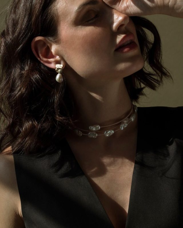 Close-up of a woman wearing a freshwater pearl drop earring with a sculptural gold ear cuff, layered with a two-strand keshi pearl and gold chain necklace, styled with a black satin top.
