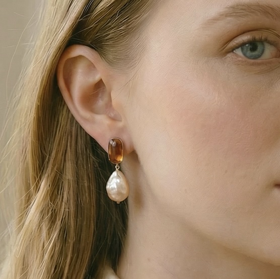Close-up of a woman's ear wearing an amber cabochon stud earring with a large baroque pearl drop in a gold setting.