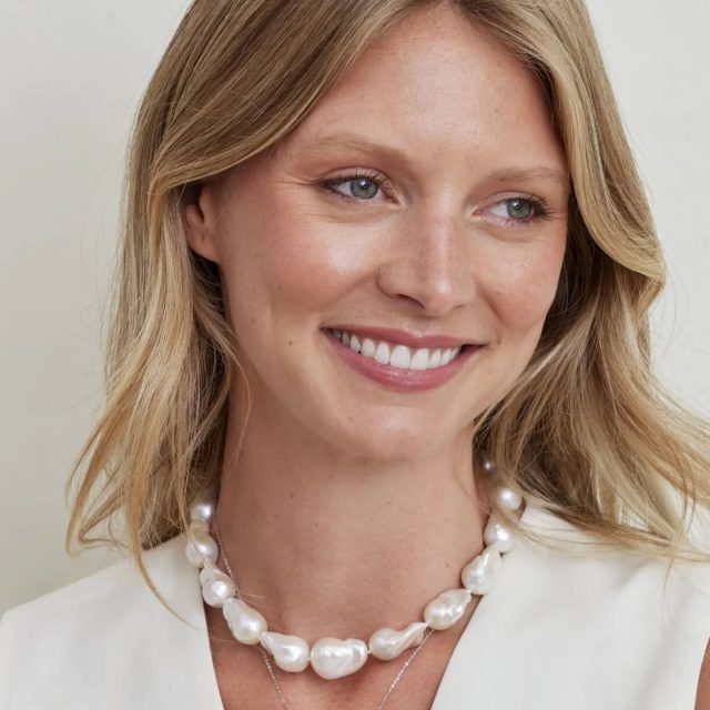 Close-up of a smiling woman wearing a large baroque freshwater pearl necklace layered over a delicate silver chain, with a white blazer.