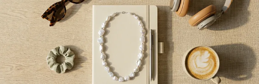 Baroque pearl necklace on a notebook beside headphones, sunglasses, and coffee.