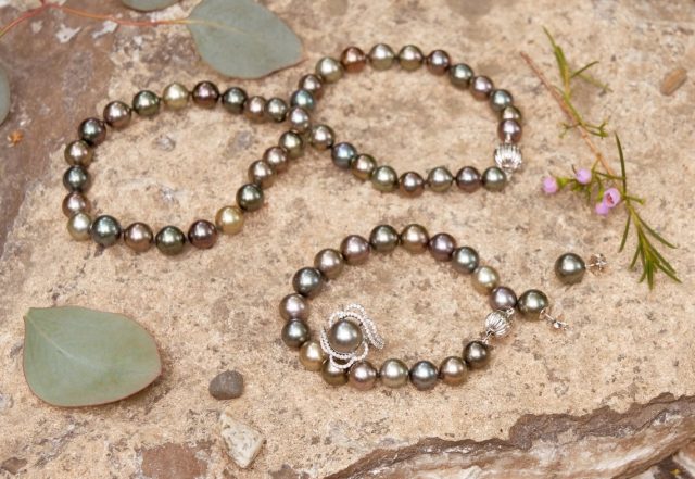 Two dark, iridescent pearl bracelets arranged on rocks with a green stone and small flowers.