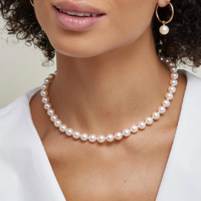 Close-up of a woman's neck and mouth wearing a classic strand of round white pearls with a gold hoop earring featuring a dangling pearl drop, worn with a white top.