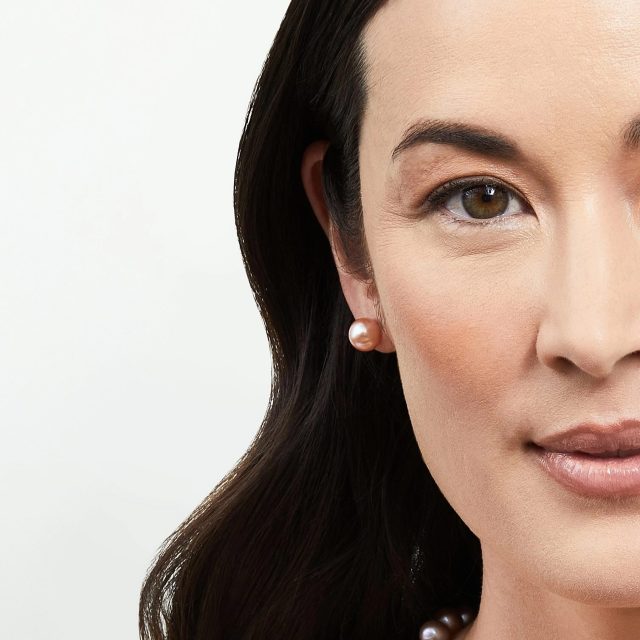 Close-up of half a woman's face wearing a single peach/pink pearl stud earring, with a hint of a dark pearl necklace visible at the collar.