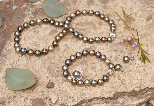 Two dark, iridescent pearl bracelets arranged on rocks with a green stone and small flowers.