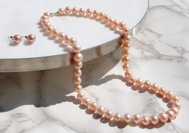 A peach pearl necklace and matching stud earrings displayed on a marble surface.