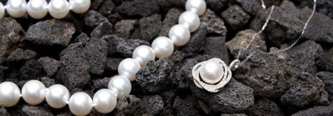 A pearl necklace, bracelet and pendant necklace displayed on dark volcanic rock, with small pink flowers in the background.