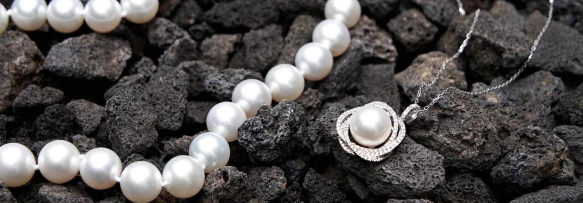 A pearl necklace, bracelet and pendant necklace displayed on dark volcanic rock, with small pink flowers in the background.