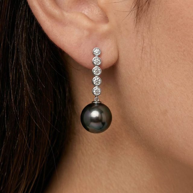 A closeup image of a woman's ear wearing a drop earring featuring five diamonds and a single black Tahitian pearl.