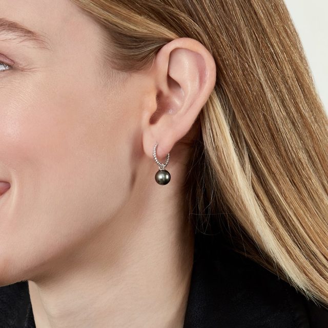 A closeup image of a woman wearing a small pair of silver hoops with a single black Tahitian pearl detail, and a black shirt.