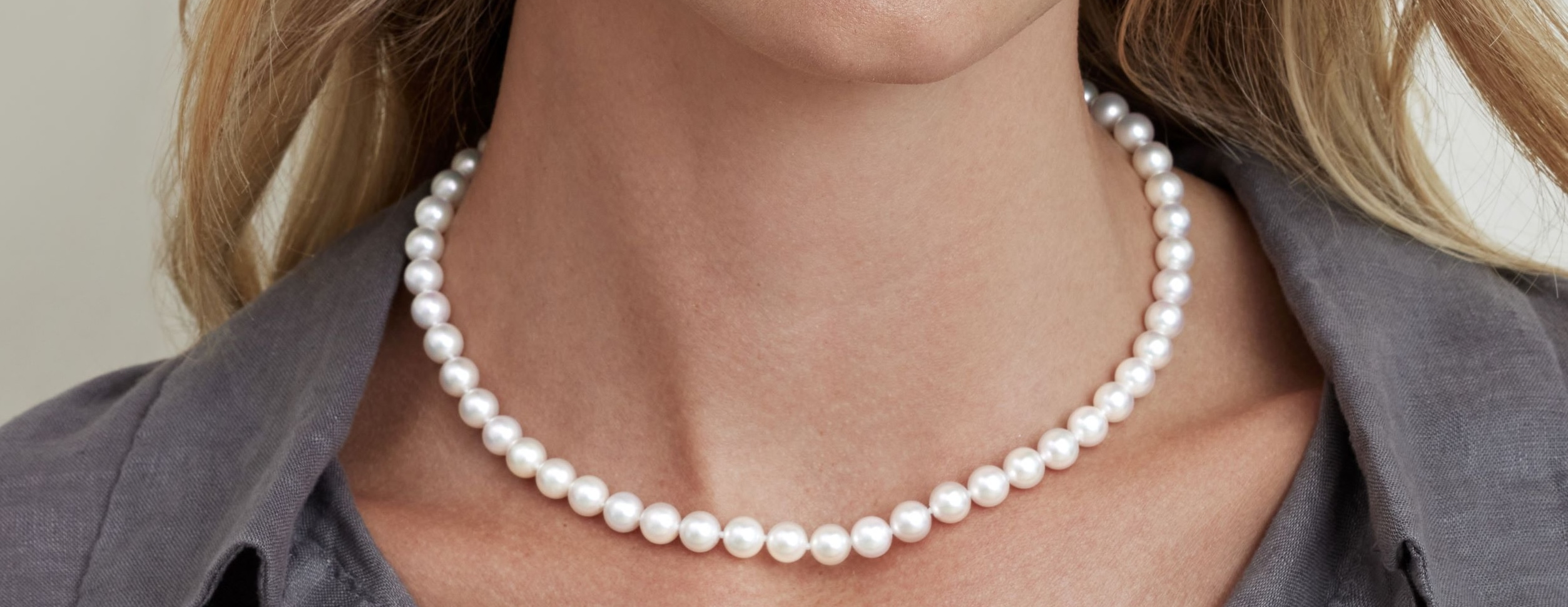 Close-up of a woman wearing a white pearl strand necklace at the collarbone, with round, closely matched pearls and a gray collared shirt.